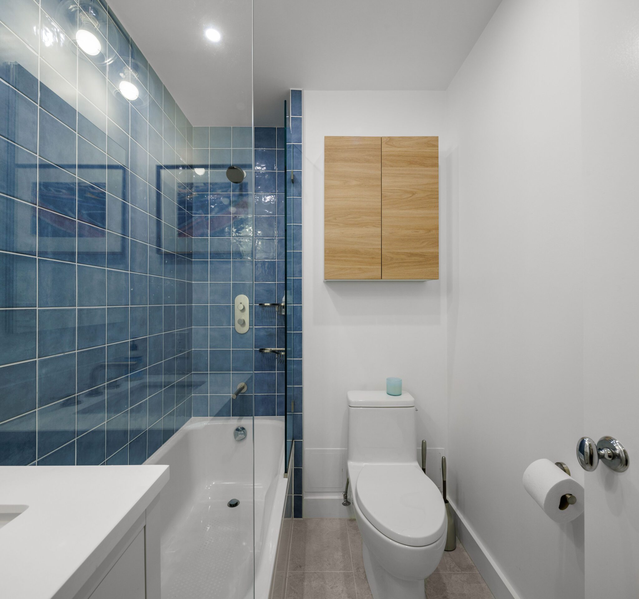 Contemporary bathroom design with blue tile shower, gold fixtures, and a wall-mounted wooden cabinet for storage.