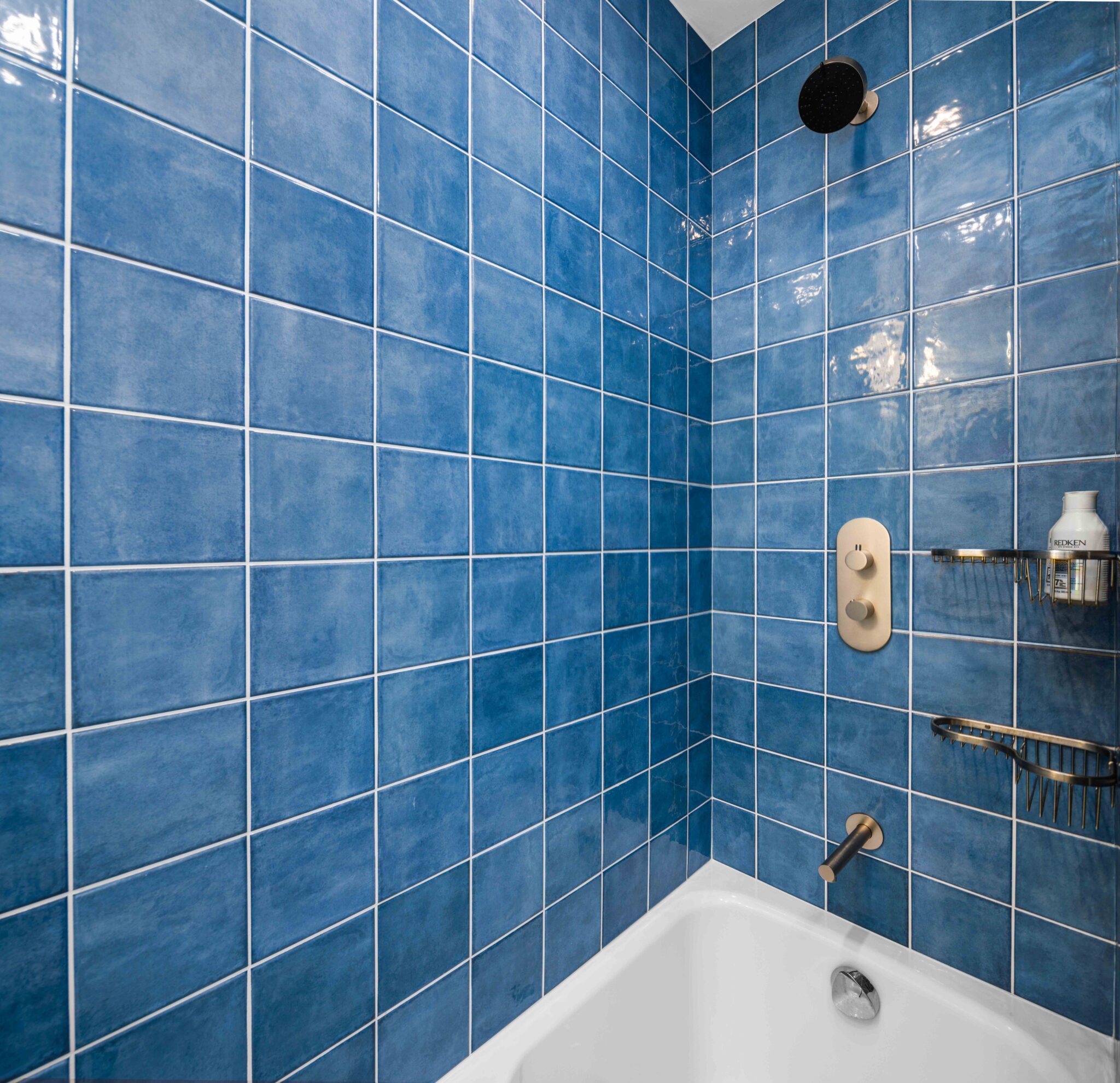 Elegant bathtub and shower combination with blue tiles and gold fixtures, featuring a rain showerhead and built-in storage.